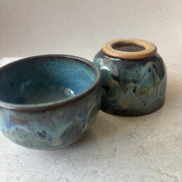 Two small handmade ceramic bowls with a glossy blue and gray marbled glaze, one standing and one lying down, set against a textured off-white background.